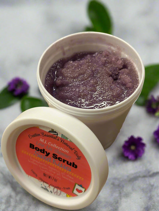 Two containers of body scrub with a purple scrub and visible label on a gray surface with green leaves.
