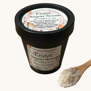 Creation Essential's Natural Body Revive creamy sugar scrub with turmeric and oats in a black jar with a wooden spoon.