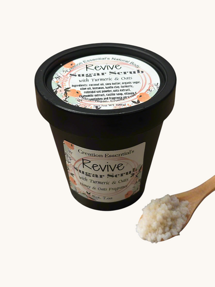 Creation Essential's Natural Body Revive creamy sugar scrub with turmeric and oats in a black jar with a wooden spoon.