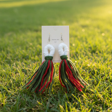 A pair of handcrafted yarn earrings with a twisted knot design, featuring colors green, red, and white, displayed on a card.
