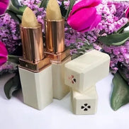 A colorless lip balm displayed alongside its packaging, which features a gold and white design. The lip balm is shown with an open lid revealing the product, and it is placed on a surface with purple flowers in the background.