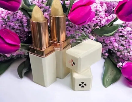 A colorless lip balm displayed alongside its packaging, which features a gold and white design. The lip balm is shown with an open lid revealing the product, and it is placed on a surface with purple flowers in the background.
