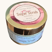 Creation Essential's Natural Body Coco Sugar Scrub in a gold-lidded jar sitting on sandy beach ground.