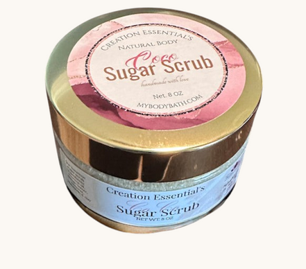 Creation Essential's Natural Body Coco Sugar Scrub in a gold-lidded jar sitting on sandy beach ground.