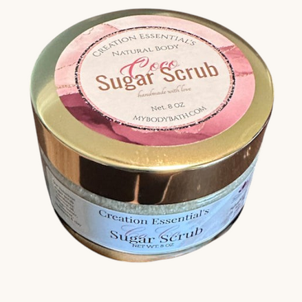 Creation Essential's Natural Body Coco Sugar Scrub in a gold-lidded jar sitting on sandy beach ground.