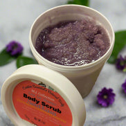 Two containers of body scrub with a purple scrub and visible label on a gray surface with green leaves.