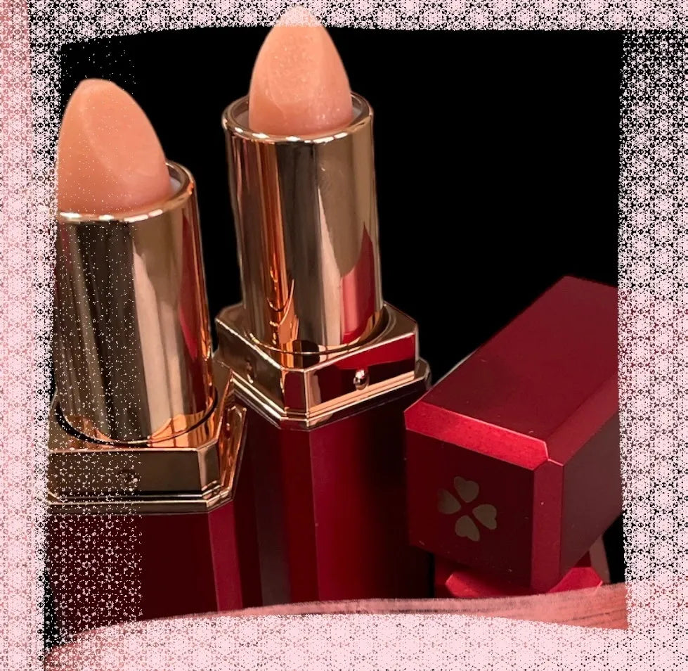 A colorless lip balm displayed alongside its packaging, which features a gold and white design. The lip balm is shown with an open lid revealing the product, and it is placed on a surface with purple flowers in the background.