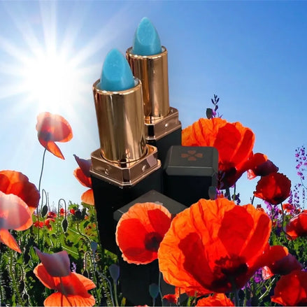 A colorless lip balm displayed alongside its packaging, which features a gold and white design. The lip balm is shown with an open lid revealing the product, and it is placed on a surface with purple flowers in the background.